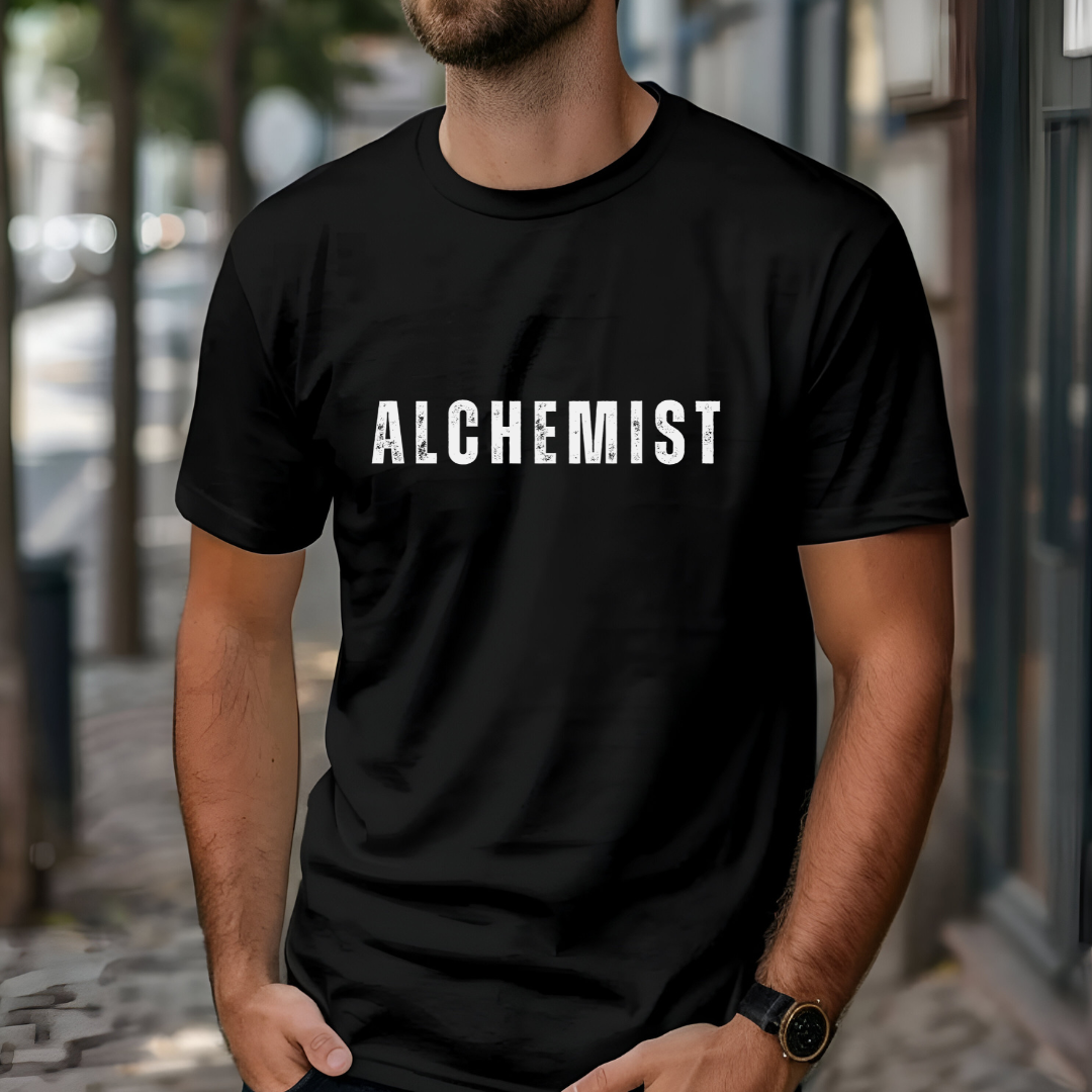 Model wearing crew neck, short sleeve T-shirt that reads 'ALCHEMIST' in distressed block letters across the chest. 'Turning Everything Into Love' in two lines of smaller block letters are written on the back. Embody love in all things.