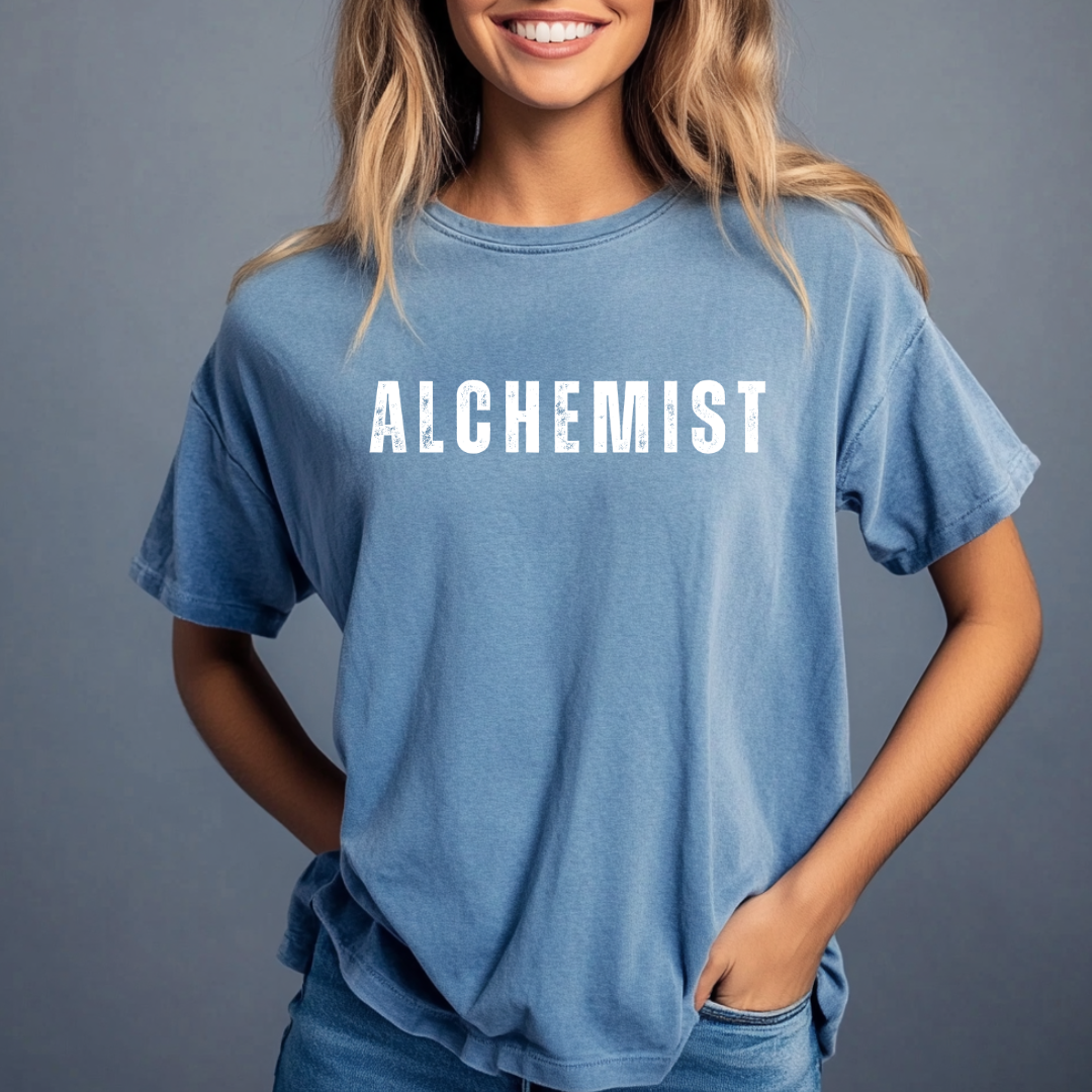 Model wearing crew neck, short sleeve T-shirt that reads 'ALCHEMIST' in distressed block letters across the chest. 'Turning Everything Into Love' in two lines of smaller block letters are written on the back. Embody love in all things.

