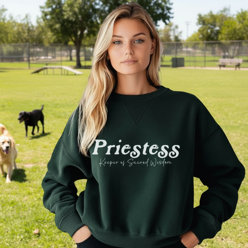 Woman wearing a green sweatshirt with 'Priestess Keeper of Sacred Wisdom' text, standing in a dog park.