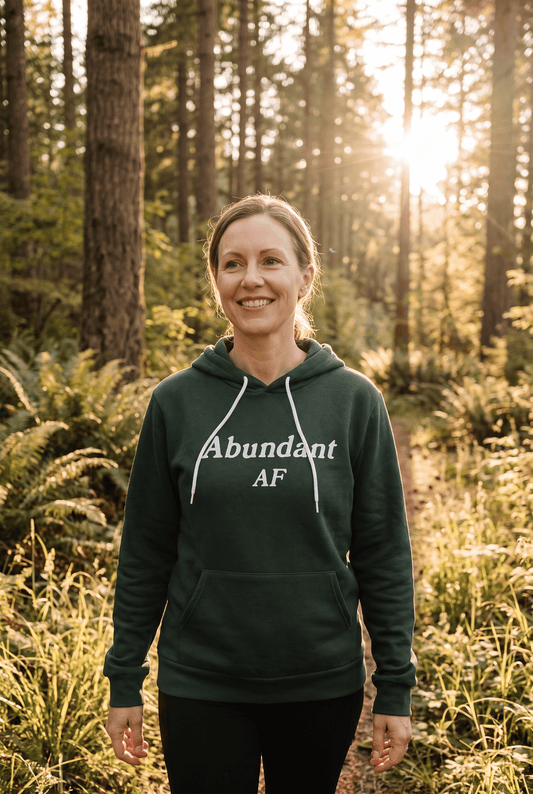 A woman in her early 40s with light skin and natural makeup walks along a golden-hour forest trail wearing the dark forest green Abundant AF hoodie. The relaxed outdoor setting reflects grounded confidence, inner peace, and an abundant lifestyle.