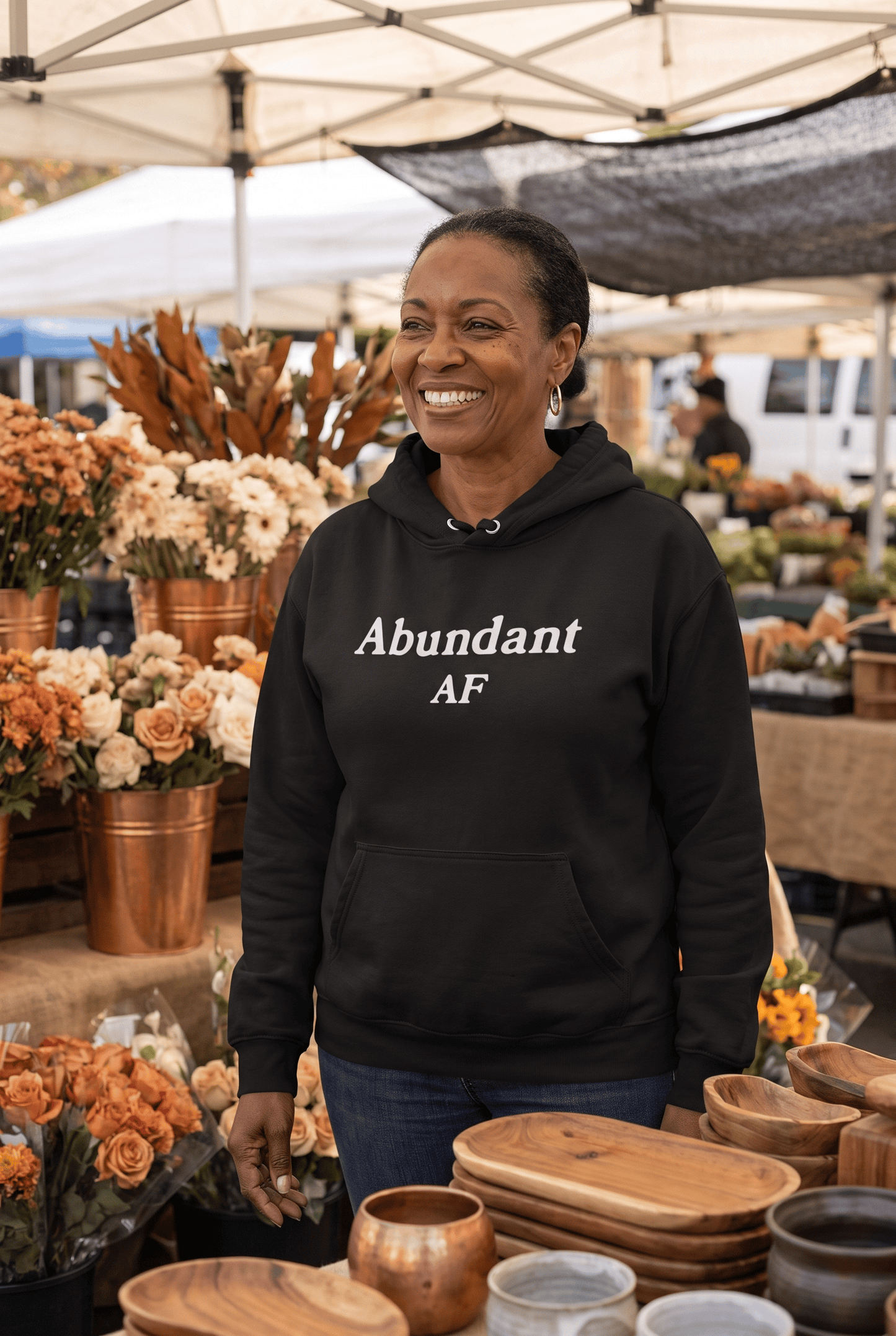 A joyful woman in her late 50s of African descent browses a charming farmer's market wearing the black Abundant AF hoodie. Warm daylight, fresh flowers, and natural textures create a welcoming atmosphere of prosperity.