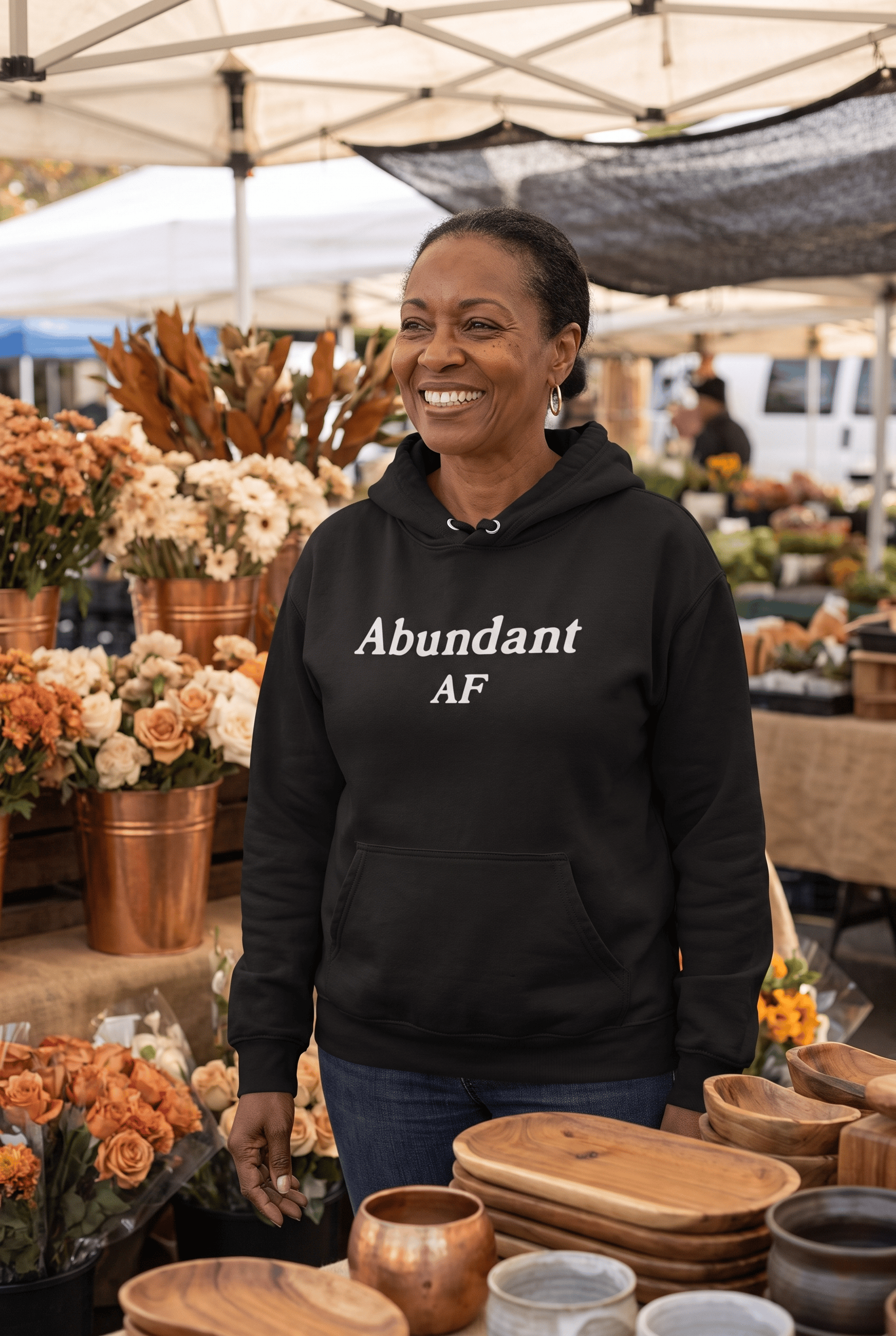 A joyful woman in her late 50s of African descent browses a charming farmer's market wearing the black Abundant AF hoodie. Warm daylight, fresh flowers, and natural textures create a welcoming atmosphere of prosperity.