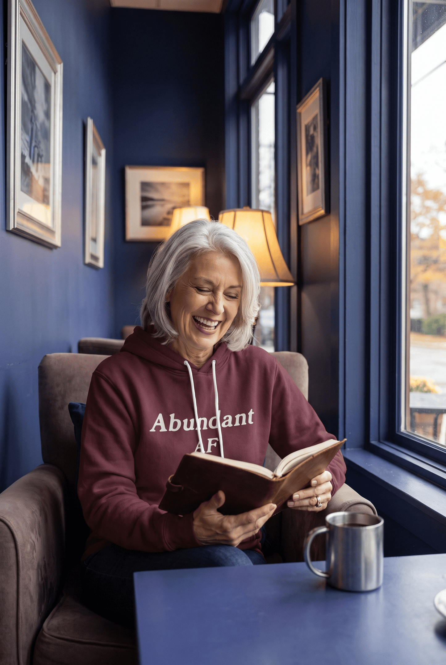 A serene woman in her early 60s with silver hair enjoys coffee beside large windows in a cozy café while wearing the burgundy Abundant AF hoodie. Warm interior lighting creates an inviting, reflective atmosphere.