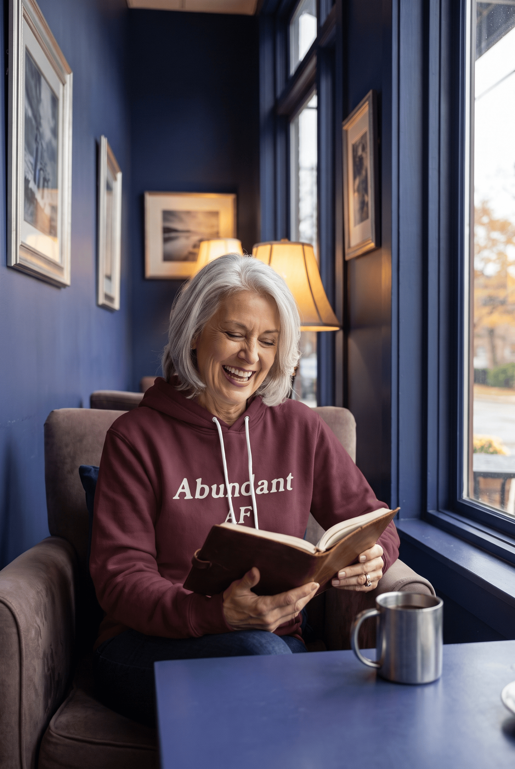 A serene woman in her early 60s with silver hair enjoys coffee beside large windows in a cozy café while wearing the burgundy Abundant AF hoodie. Warm interior lighting creates an inviting, reflective atmosphere.