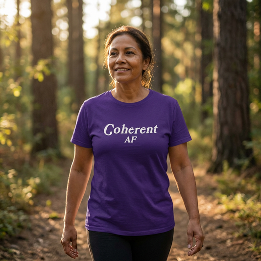 A woman in her early 50s with warm skin and soft features walks along a sunlit forest path at golden hour, wearing a purple “Coherent AF” t-shirt. The light filters through the trees, creating a calm, grounded, and empowered atmosphere. Her expression is peaceful and self-assured.