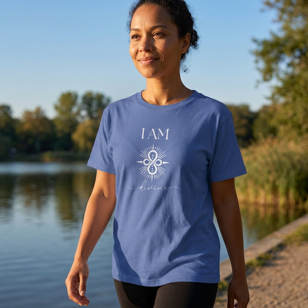 A woman walks peacefully along a lakeside path wearing a mystic blue T-shirt printed with the words “I AM Divine” and a central sigil symbol. The natural setting and soft light evoke serenity, sacred alignment, and a deep connection to inner and outer divinity.