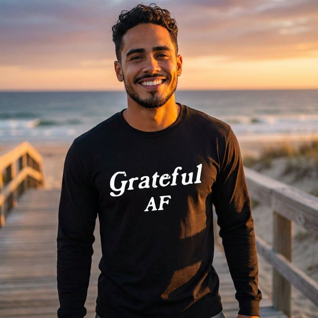 A smiling man stands on a wooden beach walkway at sunset, wearing a black long-sleeve shirt with the words “Grateful AF” in bold white lettering. The ocean and warm sky behind him evoke calm presence, joy, and the energetic embodiment of gratitude.