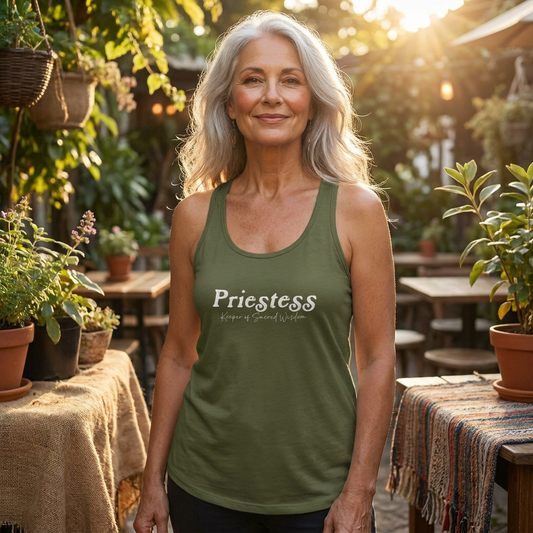 A woman in her late 50s with silver hair and a calm, radiant expression stands near a softly lit outdoor café garden wearing an olive green “Priestess, Keeper of Sacred Wisdom” racerback tank. Warm golden hour light creates a peaceful and empowered atmosphere surrounded by plants and natural textures.
