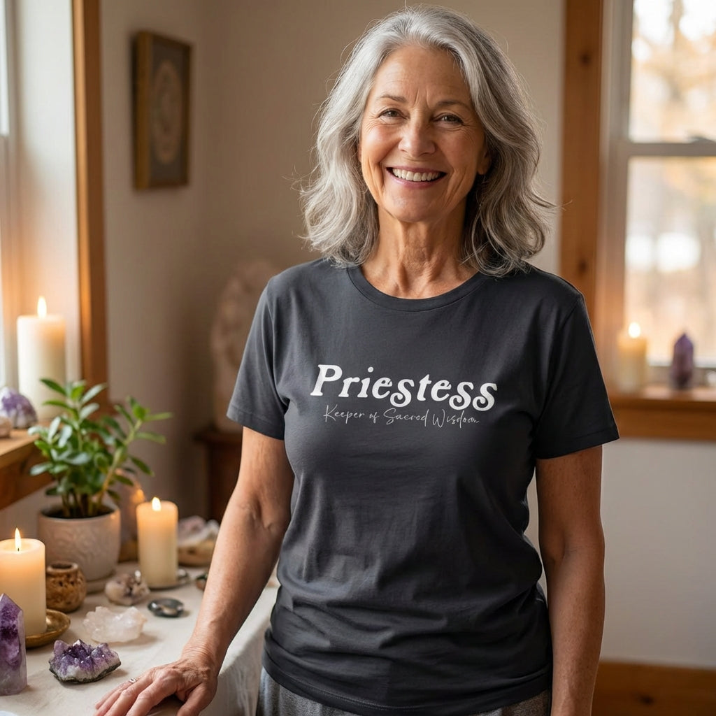 A smiling woman stands in a softly lit room with candles, crystals, and sacred objects, wearing a dark T-shirt that reads “Priestess, Keeper of Sacred Wisdom.” The setting evokes a peaceful ceremonial space, blending everyday comfort with spiritual symbolism and feminine mystique.