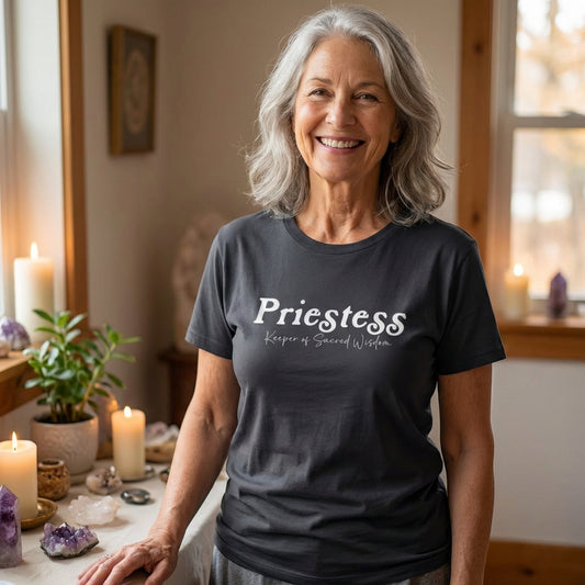 A smiling woman stands in a softly lit room with candles, crystals, and sacred objects, wearing a dark T-shirt that reads “Priestess, Keeper of Sacred Wisdom.” The setting evokes a peaceful ceremonial space, blending everyday comfort with spiritual symbolism and feminine mystique.